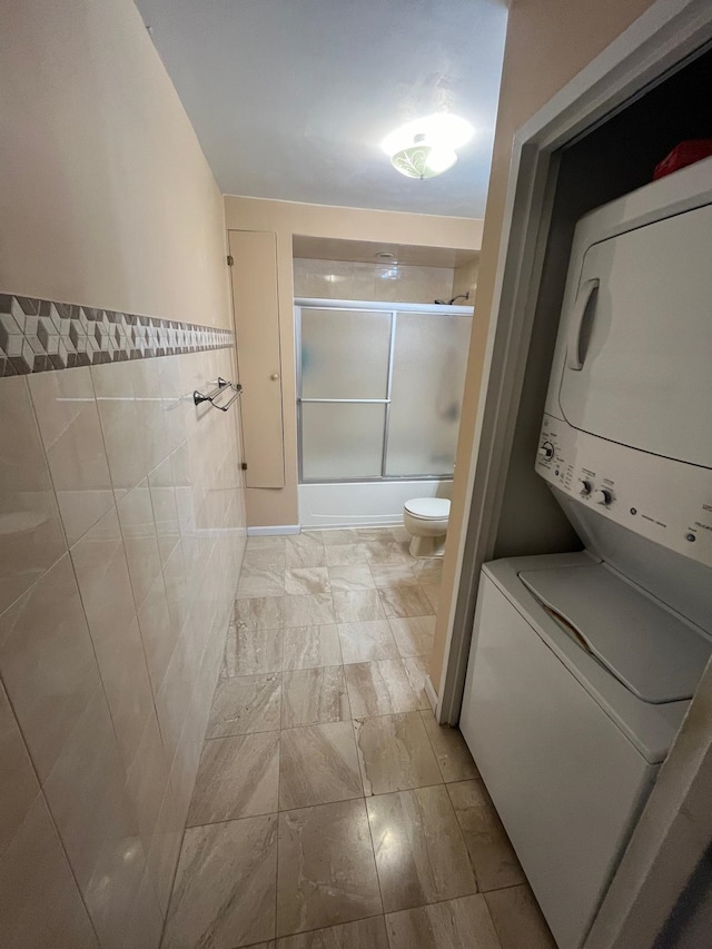 full bath with enclosed tub / shower combo, toilet, tile walls, and stacked washer and dryer