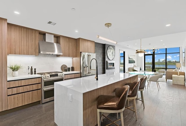 kitchen featuring a center island with sink, appliances with stainless steel finishes, a kitchen breakfast bar, dark wood-type flooring, and wall chimney exhaust hood