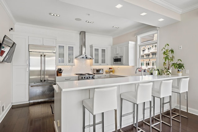 kitchen with wall chimney range hood, a breakfast bar, built in appliances, a peninsula, and glass insert cabinets