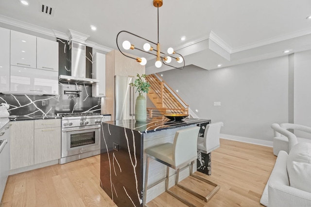 kitchen featuring wall chimney range hood, high end stainless steel range oven, hanging light fixtures, ornamental molding, and white cabinetry