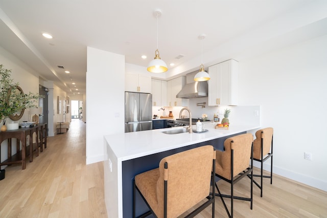 kitchen featuring a sink, stainless steel fridge, light wood-style floors, a peninsula, and wall chimney range hood