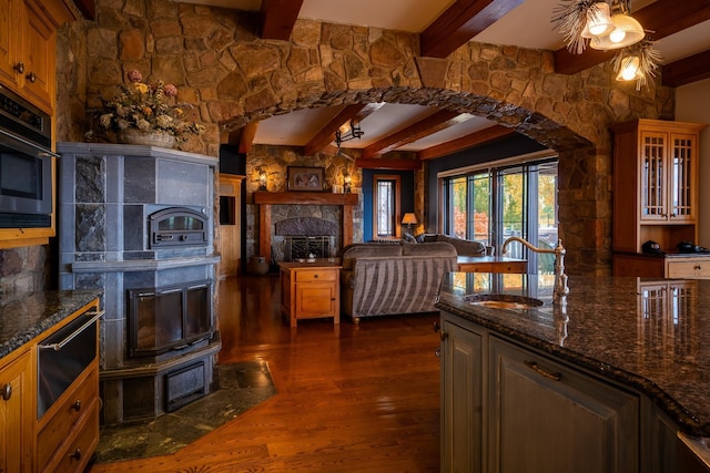 kitchen featuring hanging light fixtures, dark stone countertops, dark hardwood / wood-style flooring, beamed ceiling, and a fireplace