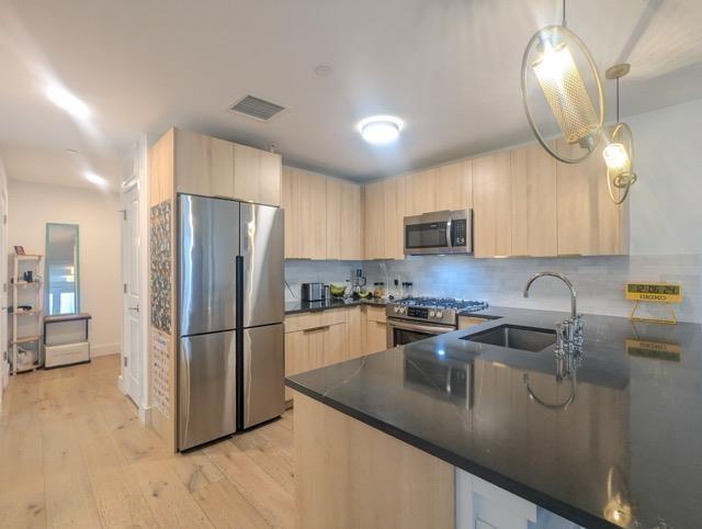 kitchen featuring light brown cabinets, a sink, appliances with stainless steel finishes, dark countertops, and pendant lighting