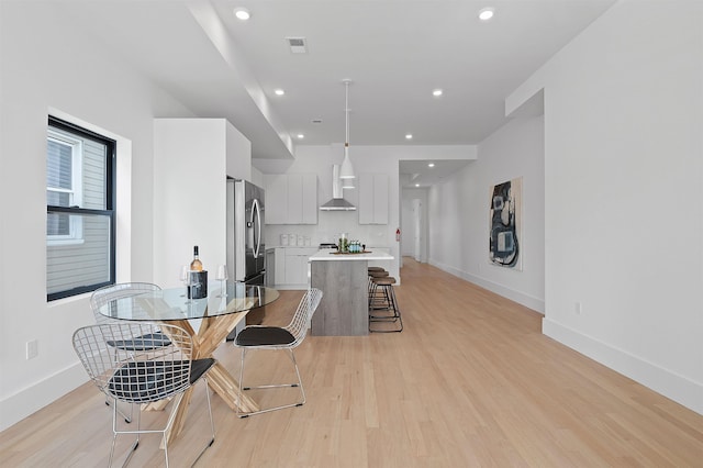 kitchen featuring decorative light fixtures, wall chimney range hood, a kitchen bar, white cabinetry, and stainless steel refrigerator