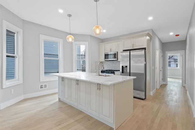 kitchen with decorative light fixtures, light stone counters, a kitchen island with sink, decorative backsplash, and stainless steel appliances