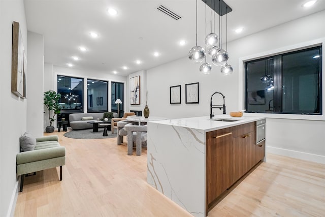 kitchen with sink, light stone counters, decorative light fixtures, a center island with sink, and light hardwood / wood-style flooring