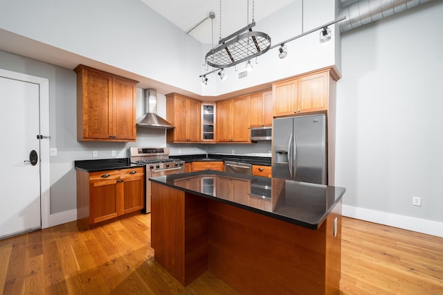 kitchen with wall chimney range hood, brown cabinets, appliances with stainless steel finishes, a high ceiling, and light wood-style floors