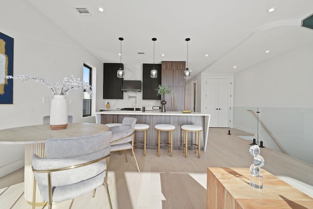 kitchen featuring decorative backsplash, hanging light fixtures, a kitchen island with sink, wall chimney range hood, and light wood-type flooring