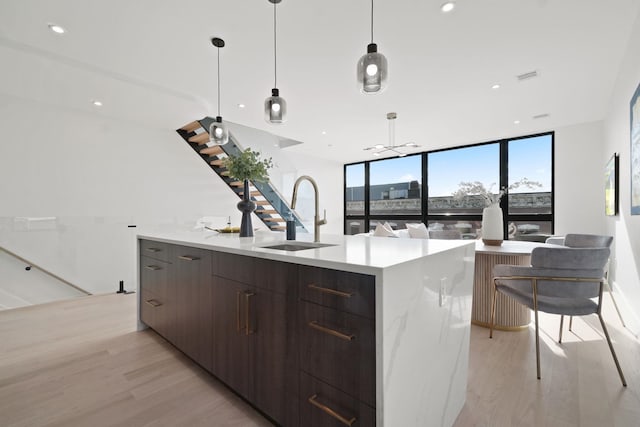 kitchen featuring sink, a kitchen island with sink, hanging light fixtures, expansive windows, and light wood-type flooring