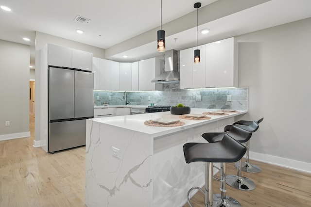 kitchen featuring white cabinets, decorative light fixtures, wall chimney range hood, and stainless steel refrigerator