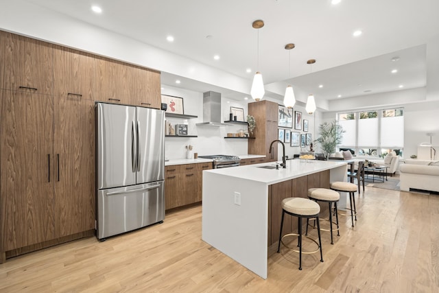 kitchen with a kitchen bar, appliances with stainless steel finishes, wall chimney exhaust hood, a kitchen island with sink, and decorative light fixtures