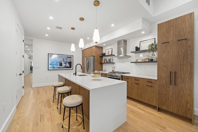 kitchen featuring stainless steel appliances, sink, wall chimney range hood, a center island with sink, and hanging light fixtures