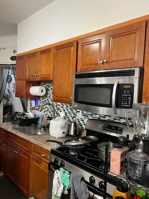 kitchen with decorative backsplash, brown cabinetry, appliances with stainless steel finishes, a sink, and light stone counters