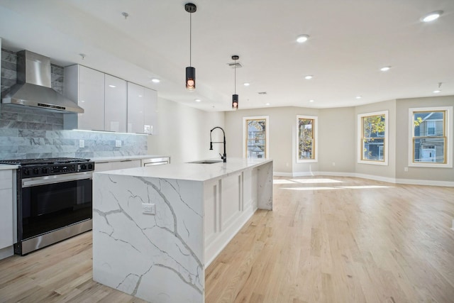 kitchen with hanging light fixtures, an island with sink, wall chimney range hood, white cabinetry, and range with gas stovetop