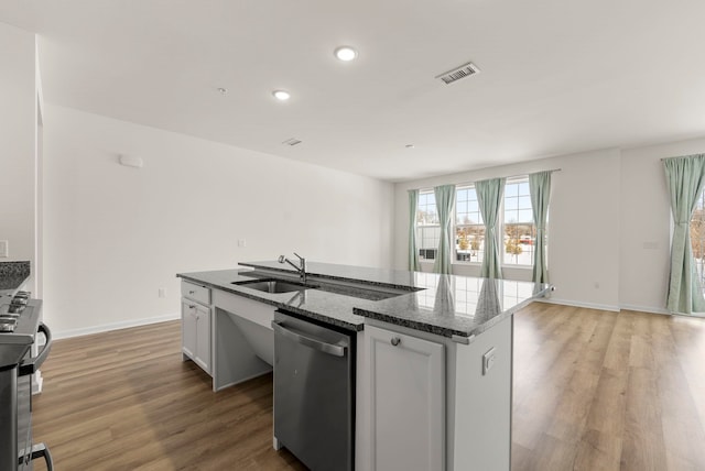 kitchen featuring appliances with stainless steel finishes, a sink, a kitchen island with sink, light wood-type flooring, and dark stone counters