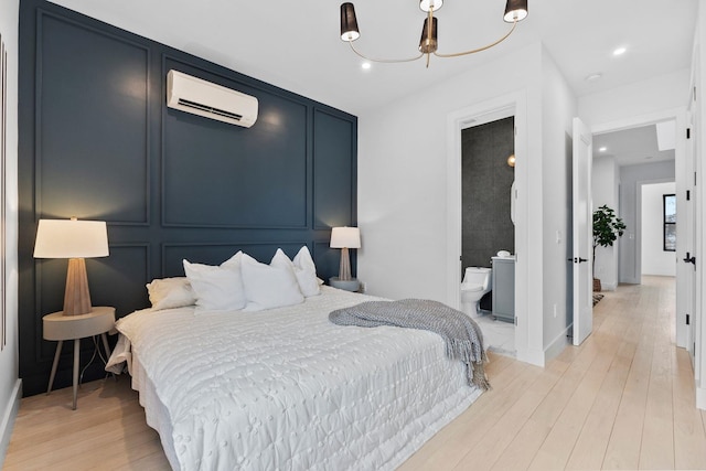 bedroom with light wood finished floors, an AC wall unit, recessed lighting, ensuite bathroom, and a decorative wall