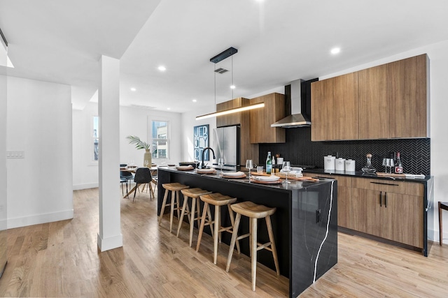 kitchen featuring dark countertops, wall chimney range hood, a kitchen island with sink, and freestanding refrigerator