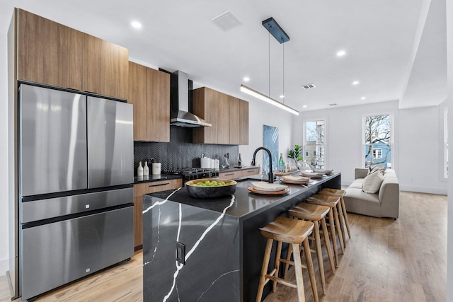 kitchen with dark countertops, an island with sink, wall chimney exhaust hood, freestanding refrigerator, and pendant lighting