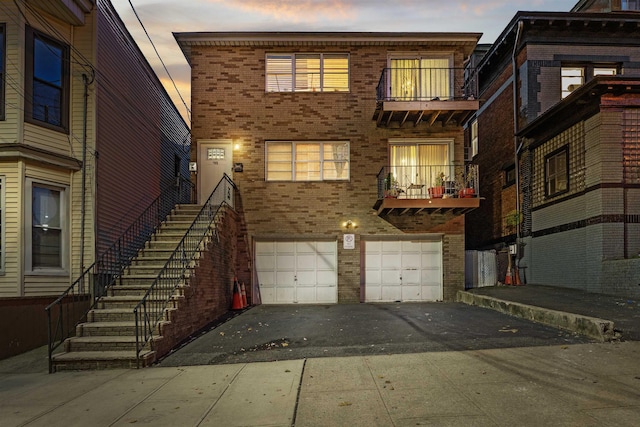 back of house at dusk with driveway, a garage, a balcony, stairway, and brick siding