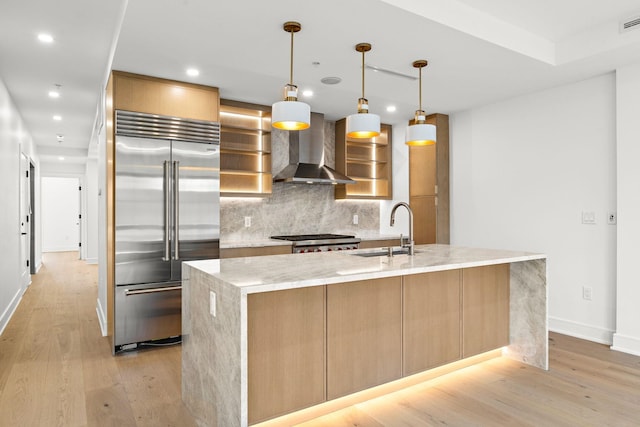 kitchen with baseboards, light wood finished floors, built in fridge, a sink, and wall chimney range hood