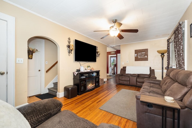 living room with arched walkways, ornamental molding, a ceiling fan, wood-type flooring, and stairs