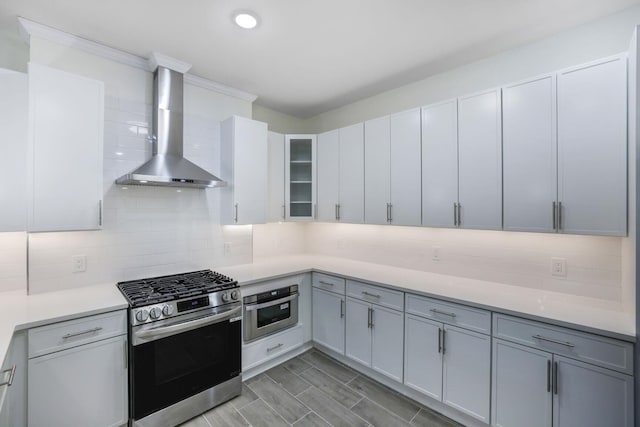 kitchen featuring stainless steel appliances, light countertops, decorative backsplash, glass insert cabinets, and wall chimney exhaust hood