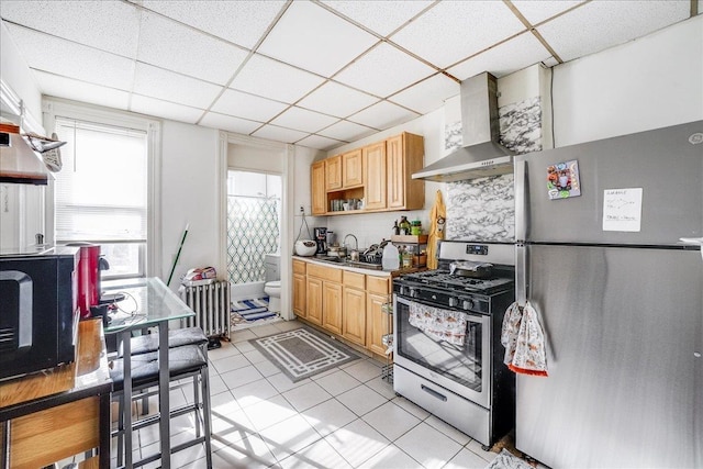 kitchen featuring a paneled ceiling, stainless steel appliances, light brown cabinetry, and wall chimney range hood