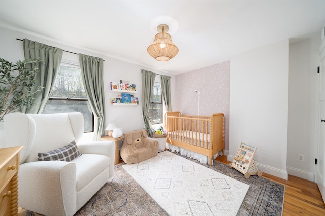bedroom featuring baseboards, wallpapered walls, a nursery area, and wood finished floors