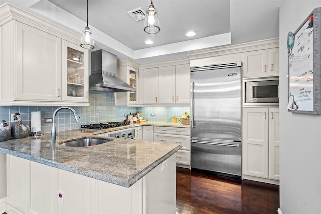 kitchen featuring wall chimney exhaust hood, a peninsula, a sink, visible vents, and built in appliances