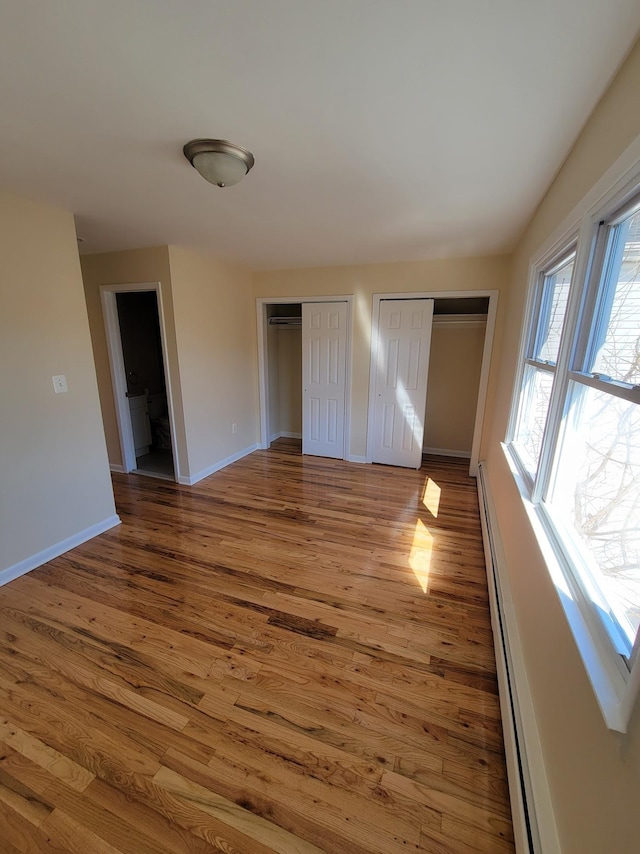 unfurnished bedroom with two closets, a baseboard heating unit, baseboards, and wood finished floors