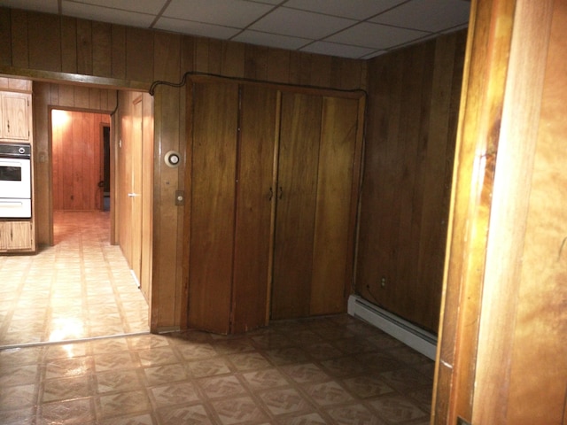 corridor featuring wooden walls, a paneled ceiling, light floors, and a baseboard radiator