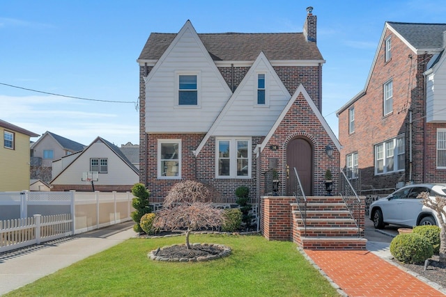 tudor home featuring fence, a front yard, a shingled roof, brick siding, and a chimney