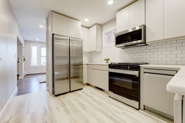 kitchen with appliances with stainless steel finishes, a baseboard radiator, white cabinets, decorative backsplash, and light wood-type flooring