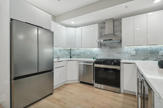 kitchen featuring light stone counters, light hardwood / wood-style flooring, appliances with stainless steel finishes, wall chimney range hood, and white cabinets