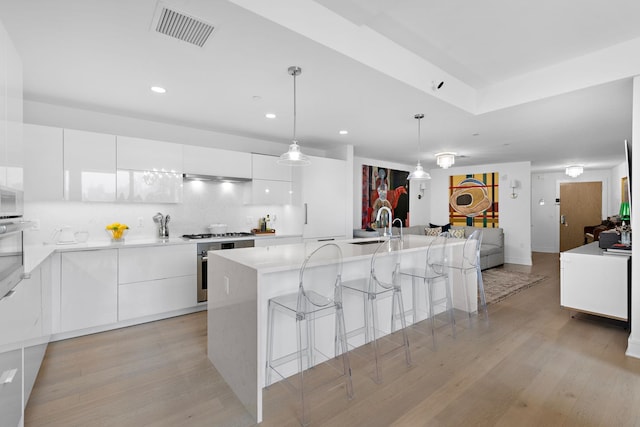 kitchen featuring tasteful backsplash, a center island with sink, light wood-type flooring, hanging light fixtures, and white cabinets