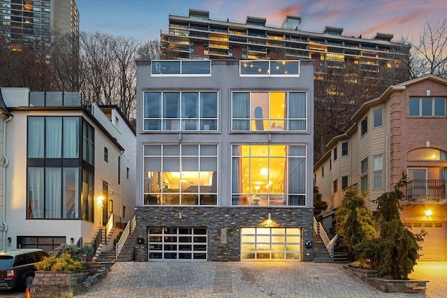 back of house at dusk with stucco siding, stairs, a garage, stone siding, and decorative driveway