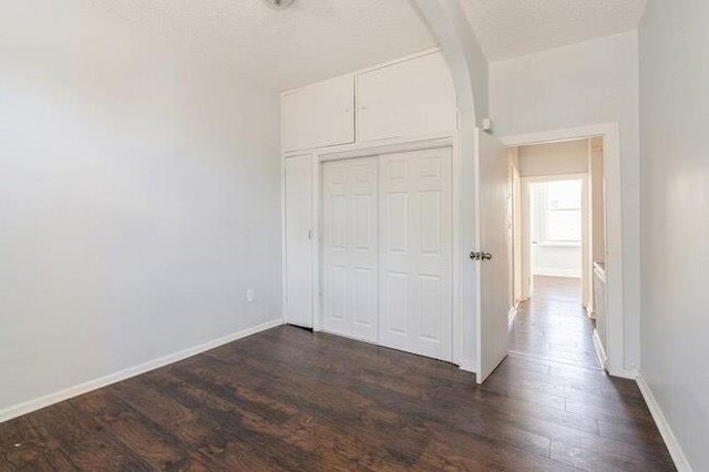 unfurnished bedroom with a textured ceiling, arched walkways, baseboards, a closet, and dark wood-type flooring