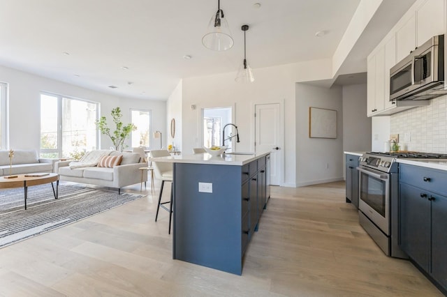 kitchen featuring tasteful backsplash, stainless steel appliances, white cabinets, hanging light fixtures, and an island with sink