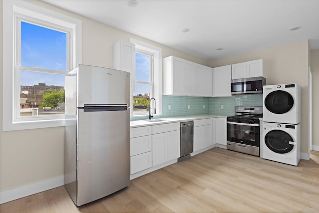 kitchen featuring stainless steel appliances, a sink, stacked washing machine and dryer, light wood-style floors, and backsplash