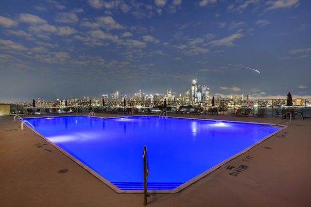 pool at twilight featuring a patio area, fence, a view of city lights, and a community pool