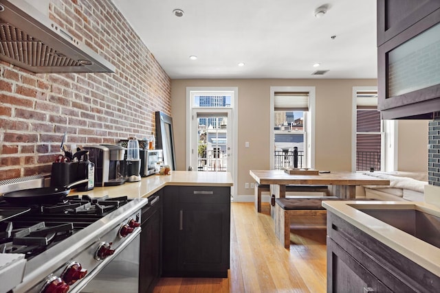 kitchen with stainless steel gas range, wall chimney exhaust hood, light hardwood / wood-style floors, dark brown cabinetry, and brick wall