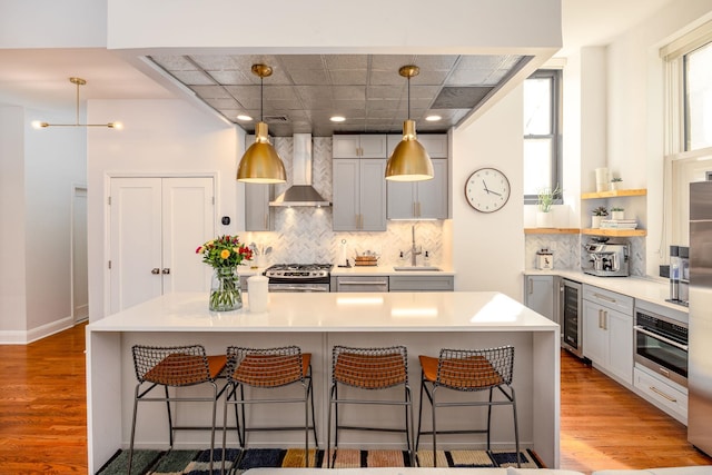 kitchen with a breakfast bar area, wine cooler, appliances with stainless steel finishes, wall chimney exhaust hood, and light wood-type flooring