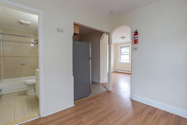 corridor featuring a baseboard heating unit, arched walkways, decorative columns, baseboards, and light wood-style floors