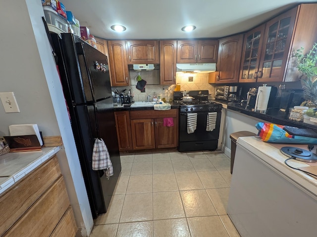 kitchen with light tile patterned floors, decorative backsplash, glass insert cabinets, under cabinet range hood, and black appliances