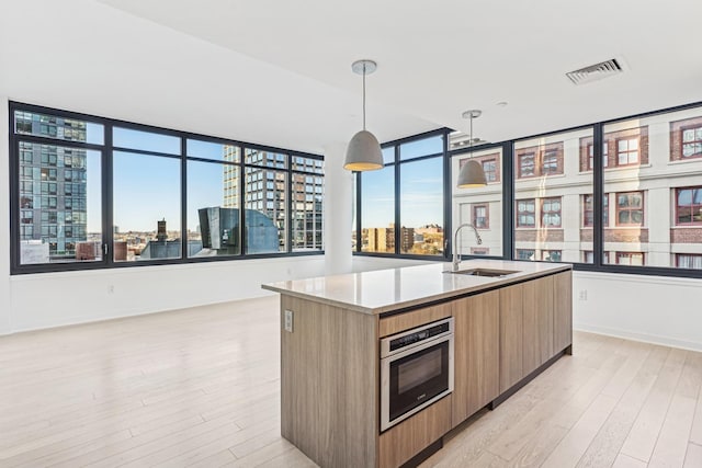 kitchen featuring stainless steel oven, sink, decorative light fixtures, light hardwood / wood-style flooring, and an island with sink