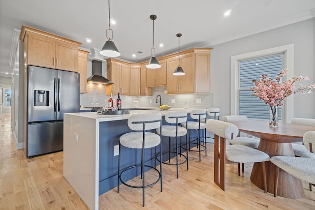 kitchen featuring wall chimney exhaust hood, stainless steel refrigerator with ice dispenser, backsplash, decorative light fixtures, and light brown cabinetry