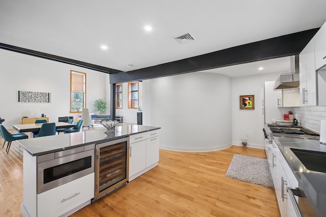 kitchen with visible vents, beverage cooler, wall chimney range hood, stainless steel microwave, and white cabinets