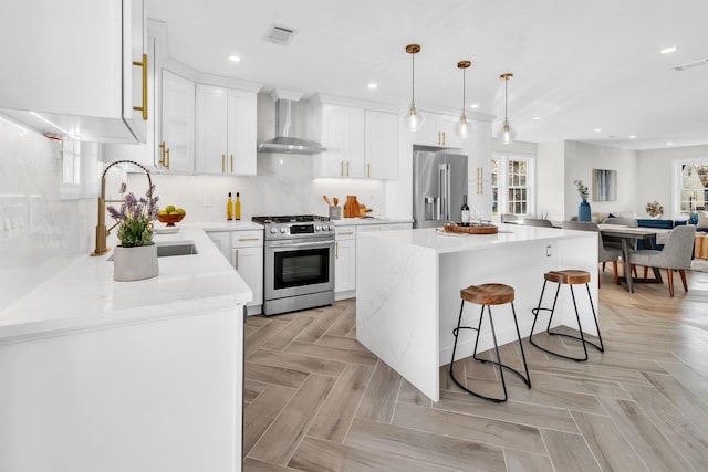 kitchen with wall chimney range hood, hanging light fixtures, appliances with stainless steel finishes, a kitchen island, and white cabinetry