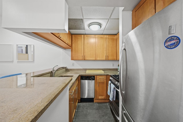 kitchen with a sink, a paneled ceiling, appliances with stainless steel finishes, dark tile patterned floors, and brown cabinets