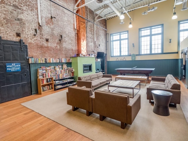 living area with wood finished floors, brick wall, a towering ceiling, wainscoting, and a glass covered fireplace